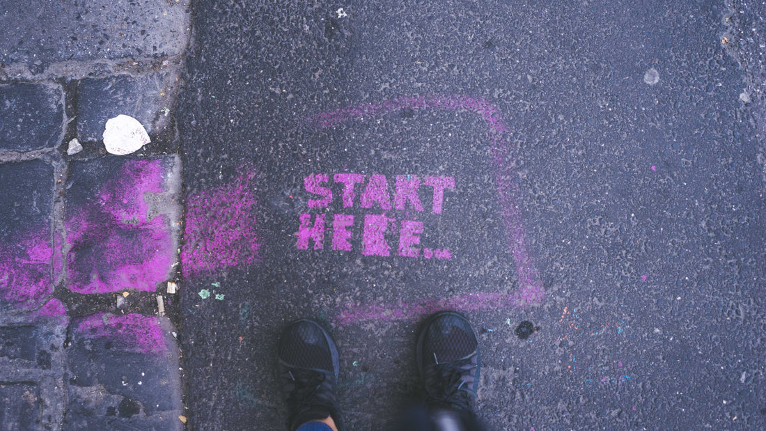 Two feet look down at graffiti on the sidewalk that says "start here", in hot pink, in contrast to the grey sidewalk.